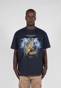 Mørkeblå bomulls t-skjorte med et grafisk motiv av en gul due med utstrakte vinger, satt mot en stormfull bakgrunn med lyn.