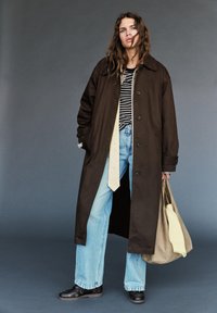 Unge kvinde iført en lang, mørk frakke, stribet skjorte, lyseblå jeans, sorte sko, holder en beige tote-taske med en sweater indeni, stående mod en grå baggrund.
