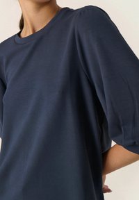 Žena v tmavě modrém topu s kulatým výstřihem a nafouklými rukávy do loktů stojí na jednoduchém světlém pozadí.