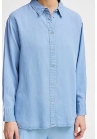 Camicia in denim azzurro chiaro con bottoni sulla parte anteriore, maniche lunghe e colletto classico. Presenta un fit rilassato e dettagli di cucitura sottili.