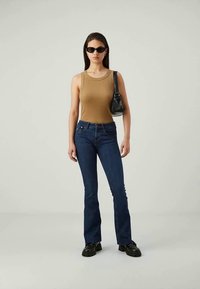 Mujer vestida con una blusa sin mangas de color beige, jeans acampanados de mezclilla azul oscuro, zapatos gruesos negros, gafas de sol negras y un pequeño bolso negro de hombro.