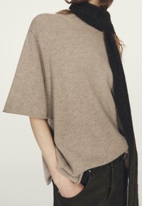 Maglione beige oversize in maglia con maniche corte, abbinato a una lunga sciarpa nera. Tessuto morbido e vestibilità rilassata. Pantaloni marrone scuro visibili.