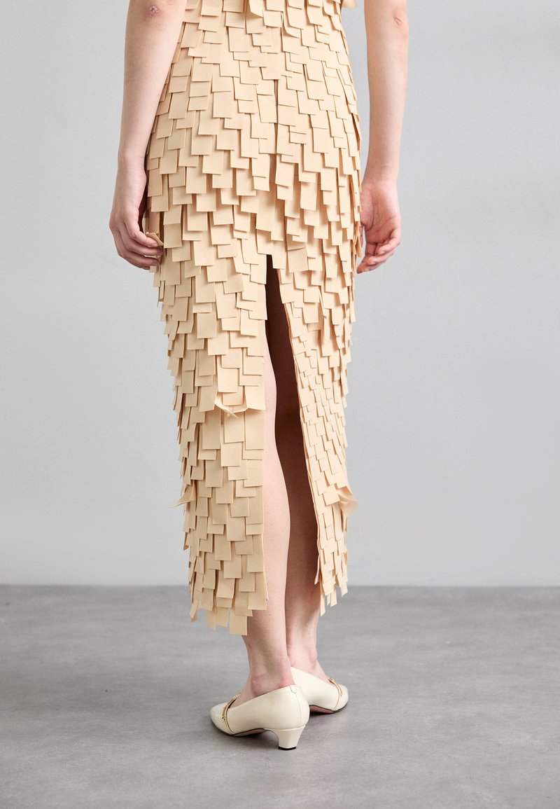 Pencil skirt in beige with a textured design of layered square fabric pieces. Features a center back slit and paired with cream-colored shoes.