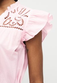 Blouse sans manches rose clair avec emmanchures volantées et broderie florale ajourée complexe sur la partie supérieure de la poitrine.