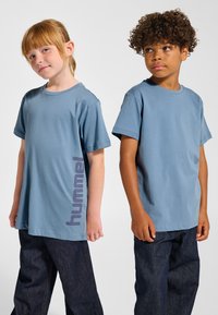 Två barn bär matchande blå kortärmade t-shirts med en "hummel"-grafik i mörklila på sidan, kombinerade med mörka denimjeans.