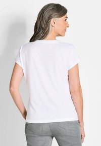 T-shirt bianco in cotone con maniche corte, scollo rotondo e vestibilità rilassata. Indossato con pantaloni in denim grigi, mostra un semplice design sul retro.