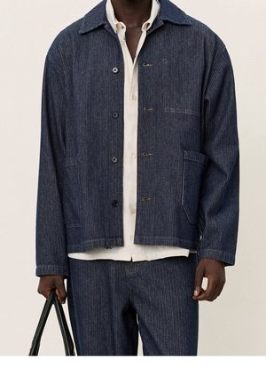 Veste bleu foncé à rayures fines en denim, avec col, fermeture par boutons et poche avant. Portée sur une chemise blanche et un pantalon.