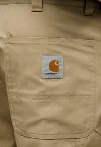 Μπεζ υφασμάτινα σορτς με πίσω τσέπη με ραμμένο περίγραμμα και τετράγωνο λογότυπο Carhartt σε πορτοκαλί και γκρι χρώμα.