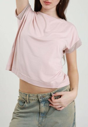 Giovane donna con un top rosa a maniche corte e jeans verdi, alza il braccio e sbottona leggermente i jeans.