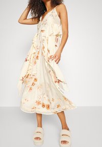 Blomstrande maxiklänning i krämfärgad med volanger, prydd med broderade detaljer och en scalloped nederkant. Modellen har på sig fluffiga vita tofflor.