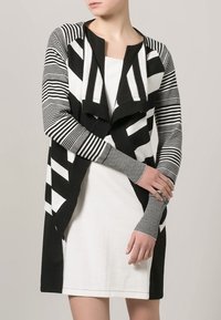 Cardigan a righe bianche e nere con apertura frontale, maniche lunghe e modelli di righe variabili; indossato sopra un vestito bianco.