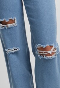 Svetlomodré deninové džínsy s dvoma veľkými poškodenými dierami na kolenách, ktoré majú rozstrapkané okraje a voľný strih po celých nohách.