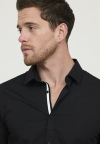 Homme avec des cheveux bruns courts et une barbe, portant une chemise noire à boutons avec un liseré blanc le long de la ligne des boutons, regardant vers la gauche.