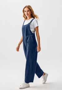 Marineblauer Jumpsuit mit weitem Bein, verstellbaren Trägern und Vordertaschen, kombiniert mit einem weißen T-Shirt und weißen Sneakers. Glatte Textur.