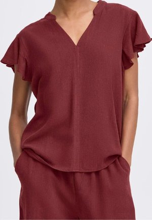 Kvinde iført en løs, kortærmet V-hals top i vinrød med flæseærmer og matchende vinrøde bukser, hænderne i lommerne.