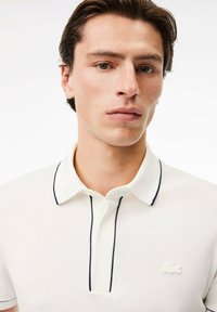 Vit polotröja med marinblå detaljer, med en tvåknapps knäppning, korta ärmar och broderad logotyp på bröstet. Strukturerad tyg.