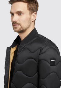 Black quilted puffer jacket with ribbed collar, tan lining, and a black logo patch. Features wave-shaped stitching and a zipper front.