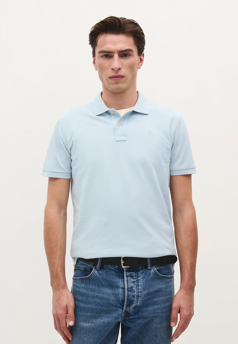 Lyseblå polo shirt med klassisk krave, korte ærmer og to knapper. Bæres med mørkeblå jeans og et sort bælte.