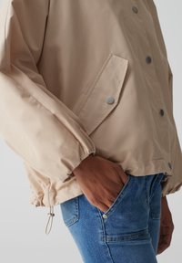 Giacca leggera beige con vestibilità rilassata, polsini elasticizzati, bottoni a scatto e tasca laterale. Indossata sopra dei jeans di denim blu.