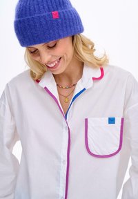 Bílá košile na knoflíky s růžovým a modrým lemem, s kapsou s modrým štítkem. Celkový vzhled doplňuje měkká fialová pletená čepice.