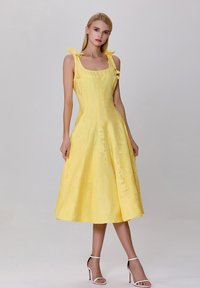Robe jaune avec un corsage ajusté et une jupe évasée, dotée de bretelles à nouer et d'une surface texturée, complétée par des sandales à talons blancs.