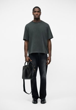 Joven de pie con vaqueros negros desgastados y camiseta gris oscuro de gran tamaño, sosteniendo un maletín de cuero negro con correa para el hombro.