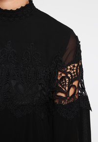 Černé šaty s vysokým krajkovým límcem, zdobené složitým krajkovým detailem na rukávech a průhlednou textilií navrch. Texturovaný a elegantní design.