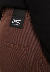 Brązowe i czarne spodnie w paski z czarną metką z logo nożyczek oraz napisem "DENHAM" blisko pasa, z widocznymi detalami szwów.