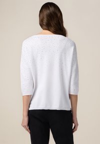 Maglione bianco con maniche a tre quarti, caratterizzato da una texture di borchie d'argento sparse sulla parte posteriore. Tessuto a maglia morbido, vestibilità rilassata.