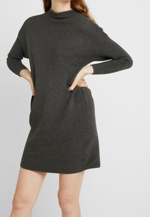 Frau trägt ein dunkelgraues, geripptes Strickkleid mit langen Ärmeln, die Hände in die Hüften gestützt, vor einem einfarbigen hellen Hintergrund.
