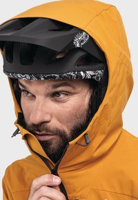 Mann mit einem schwarzen Fahrradhelm mit weißen Mustern und einer senfgelben Kapuzenjacke, der den Kragen der Jacke hochzieht.