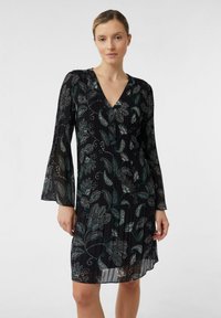 Frau in einem schwarzen langärmligen Kleid mit blau-weißen Blumenmustern, die vor einem schlichten weißen Hintergrund steht.