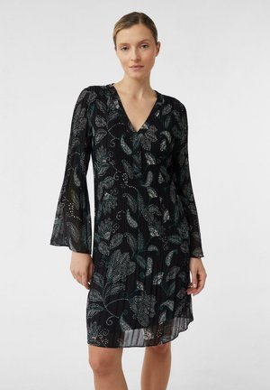 Frau in einem schwarzen langärmligen Kleid mit blau-weißen Blumenmustern, die vor einem schlichten weißen Hintergrund steht.