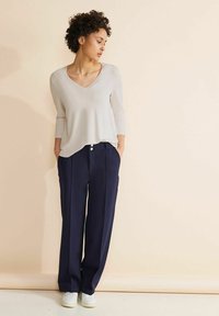 Maglione beige chiaro a scollo a v con maniche lunghe abbinato a pantaloni larghi blu navy con dettaglio a bottone in vita e sneakers bianche.