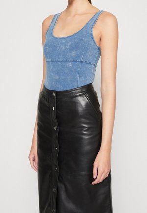 Kobieta ubrana w bezrękawnik z wyblakłego niebieskiego denimu, zestawiony z wysokim stanem, czarną spódnicą skórzaną zapinaną na guziki, na białym tle.