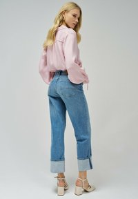 Camisa rosa clara de manga larga con corte holgado y detalle de lazo en la cintura, combinada con jeans de mezclilla azul claro de talle alto con dobladillos remangados.