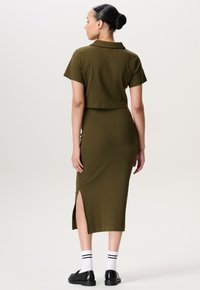 Vrouw met donker haar in een bun, gekleed in een olijfgroene geribde top met korte mouwen en een bijpassende midi-rok met een zijsplit, witte sokken en zwarte instappers, met de rug naar de camera gericht.