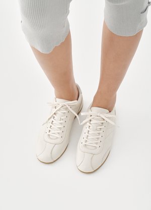 Άσπρα αθλητικά παπούτσια από δέρμα με υφή, στρογγυλή μύτη και επίπεδα κορδόνια. Η εξωτερική σόλα είναι από καουτσούκ για καλύτερη πρόσφυση. Φοριούνται με γκρι παντελόνι cropped.