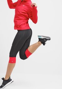 Persona con chaqueta roja y mallas negras en medio de un salto, con una pierna doblada, usando zapatillas de correr negras, sobre un fondo blanco liso.