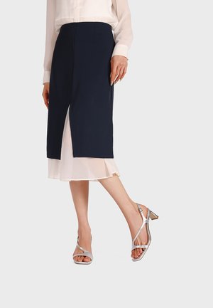 Donna che indossa una gonna blu navy al ginocchio con spacco frontale sopra un vestito rosa chiaro, abbinata a sandali argento con tacco basso e punta aperta.