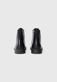 Botas de tobillo de cuero negro con textura suave, cierre con cremallera trasera y suela de goma negra en contraste. Diseño minimalista.