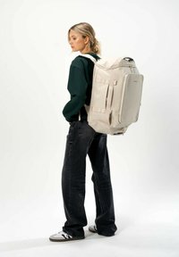 Sac à dos beige avec des bretelles rembourrées, des fermetures à glissière et des boucles latérales. Présente une texture lisse et un design rectangulaire structuré.