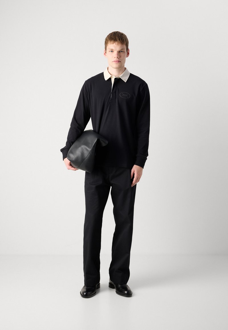 Zwarte lange mouwen polo shirt met een crèmekleurige kraag, een ritssluiting aan de voorkant en een logo patch, gecombineerd met zwarte broeken en een zwart leren tas.
