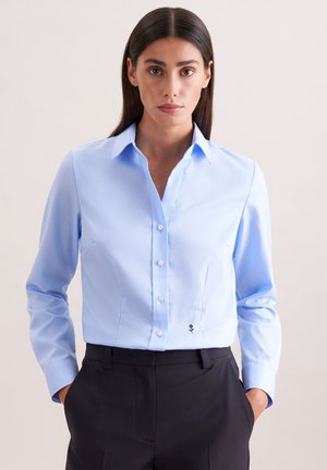 Femme aux longs cheveux bruns portant une chemise bleu clair boutonnée rentrée dans un pantalon noir taille haute, debout les mains dans les poches.