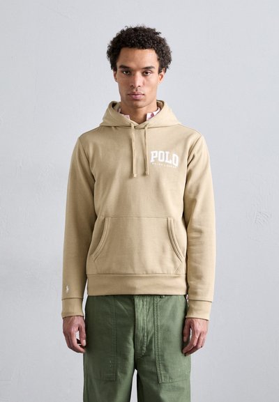 Lys beige hoodie med en frontkangaroo-lomme, snoretræk-hætte og hvid "POLO" logo. Sammen med grønne cargo-bukser. Materialet er en blanding af bomuld.