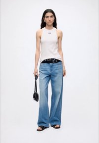 Jeune femme aux longs cheveux foncés portant un haut blanc sans manches, un jean bleu à jambes larges, des sandales noires et tenant un petit sac à main noir.