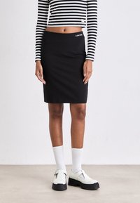 Crna olovka suknja s Calvin Klein logotipom na pojasu, u paru s crno-bijelom prugastom kročenom majicom i punim bijelim cipelama.