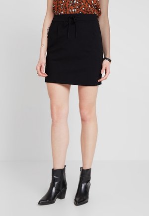 Mini jupe noire avec une ceinture à cordon, fabriquée dans un tissu lisse. Associée à des bottines noires.