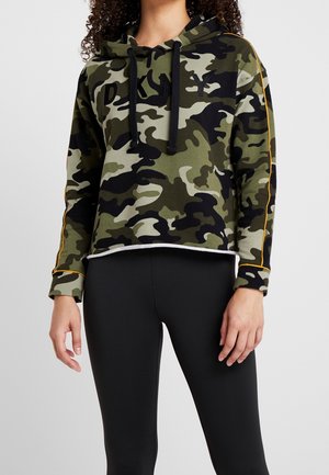 Personne portant un sweat à capuche camouflage vert et noir avec des cordons noirs et des leggings noirs sur un fond clair uni.
