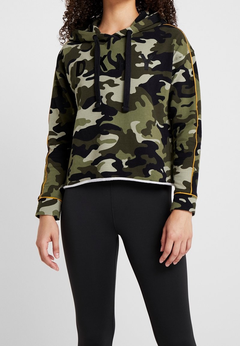 Personne portant un sweat à capuche camouflage vert et noir avec des cordons noirs et des leggings noirs sur un fond clair uni.